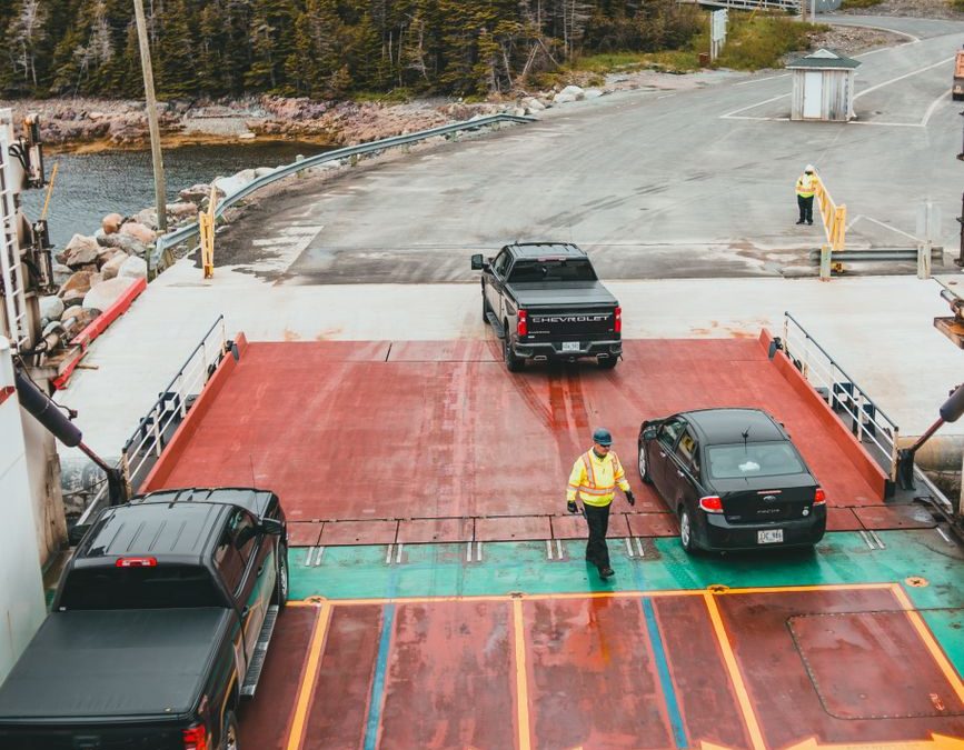 Trucks and cars exit a ferry, guided by workers at a dock in a rural setting.