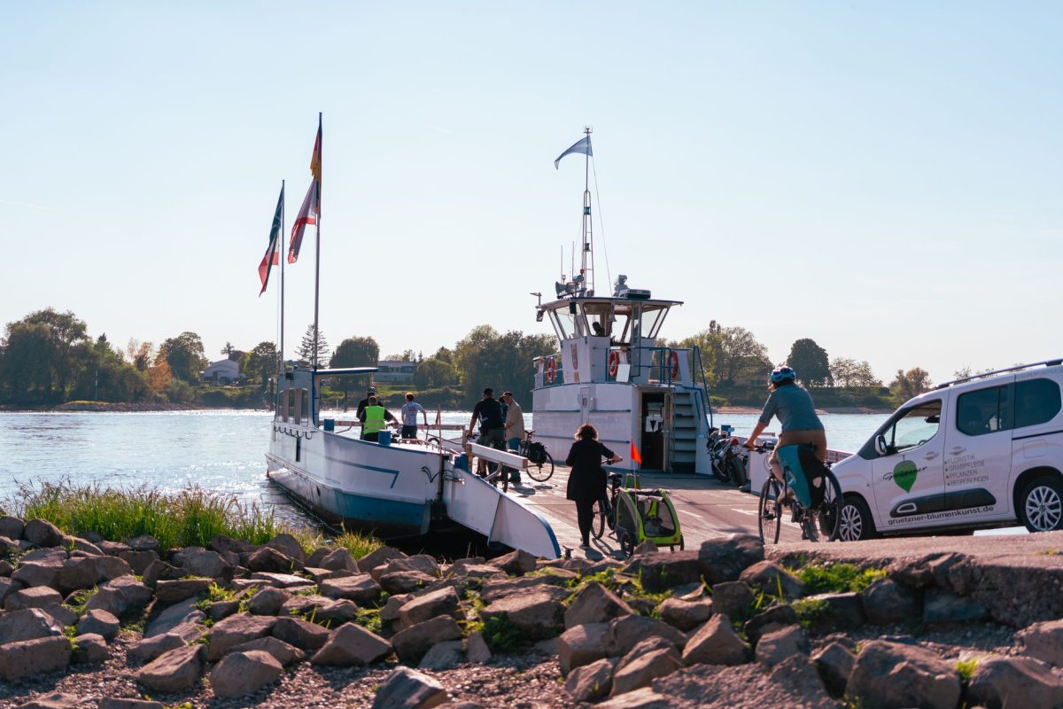 People with bicycles board a ferry on a calm river under a clear blue sky.