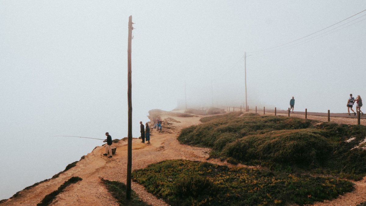 People walking and fishing along a foggy coastal path in Nazaré, Portugal.