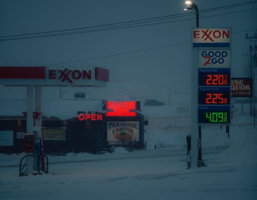 A snowy winter scene of an Exxon gas station with illuminated price signs at dusk.
