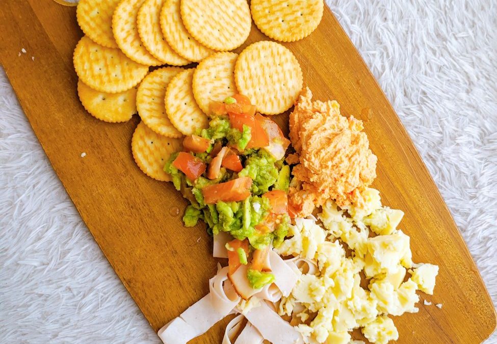 Top view of a snack platter with crackers, ham, scrambled eggs, and guacamole on a wooden board.