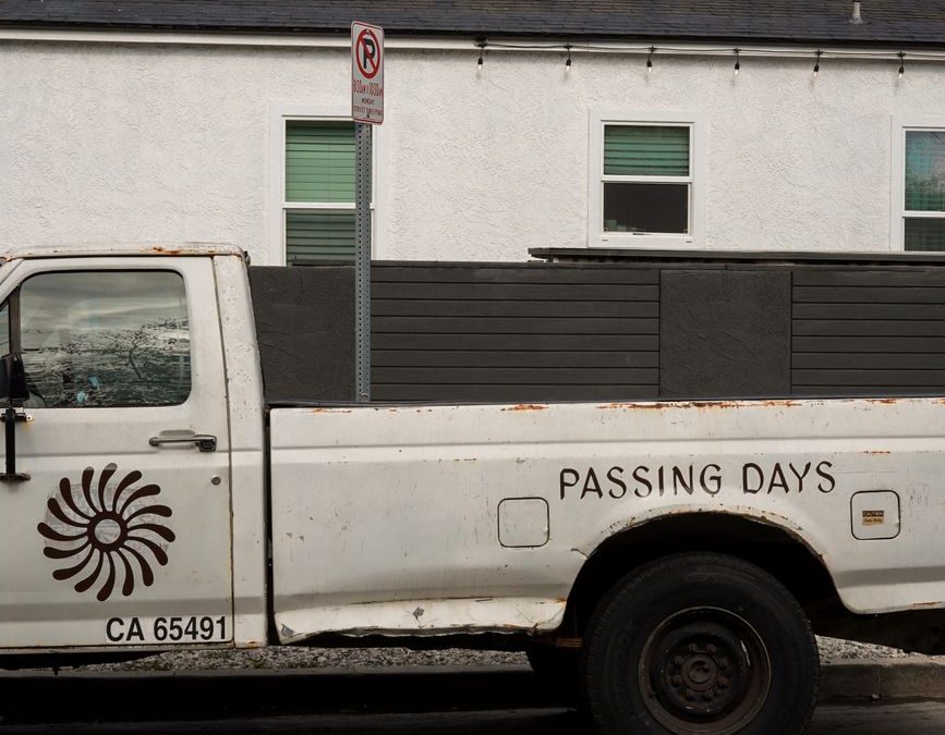 A rusty pickup truck with &amp;#39;Passing Days&amp;#39; parked on a city street against a residential building.