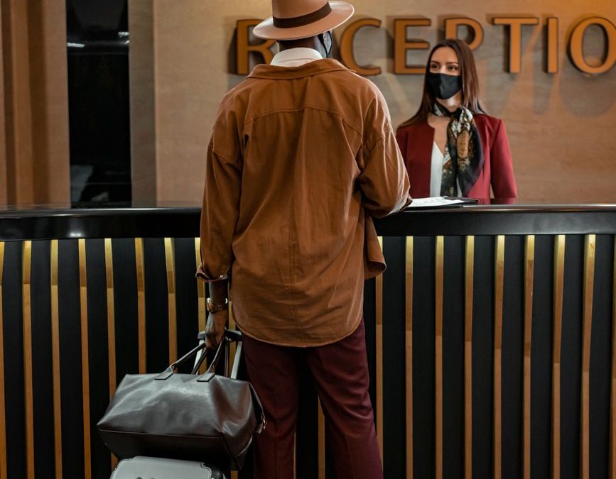 5 Hotel Habits That Make Travelers Seem Unprepared 1 Man with luggage checking in at hotel reception. Staff wearing mask, indoors, travel theme.