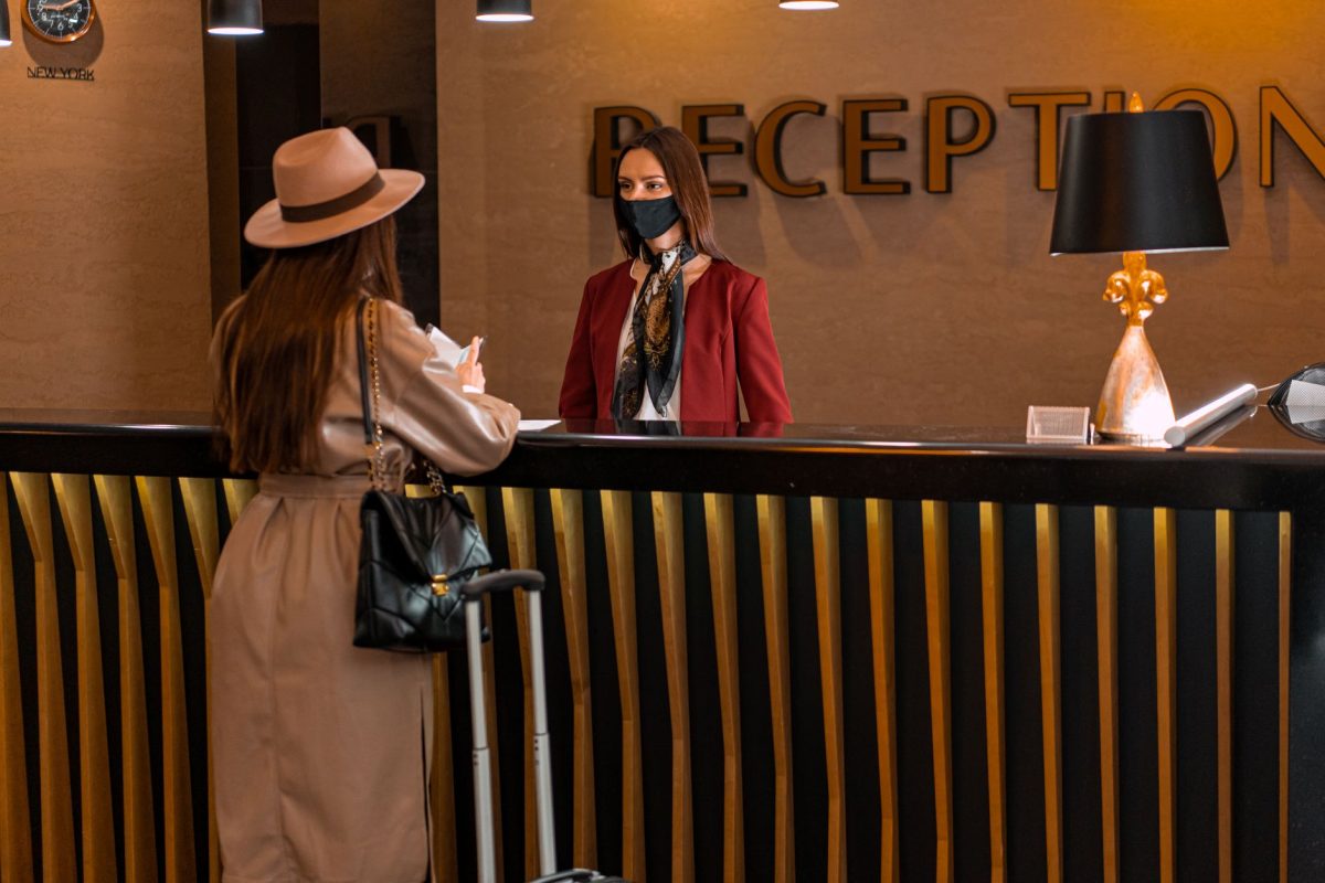 5 Hotel Habits That Make Travelers Seem Unprepared 4 A woman speaks to a receptionist wearing a mask at a hotel reception.