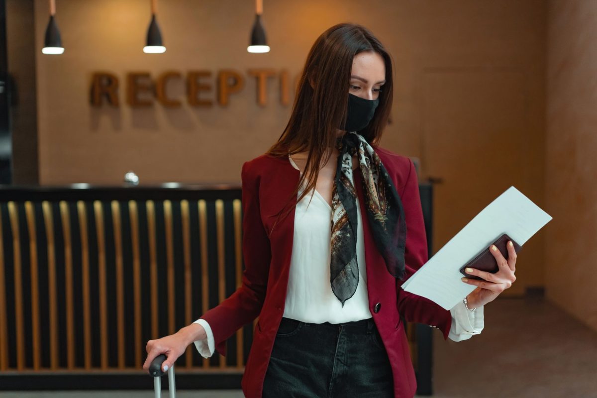 5 Hotel Habits That Make Travelers Seem Unprepared 5 Woman in red blazer at hotel reception, holding luggage and documents, wearing a face mask.