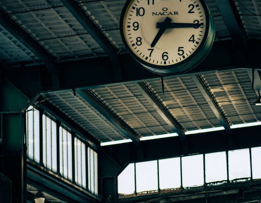5 Train Station Mistakes That Make Trips Go Sideways Fast 2 Large clock hanging at an industrial train station with metal roofing, indicating time.