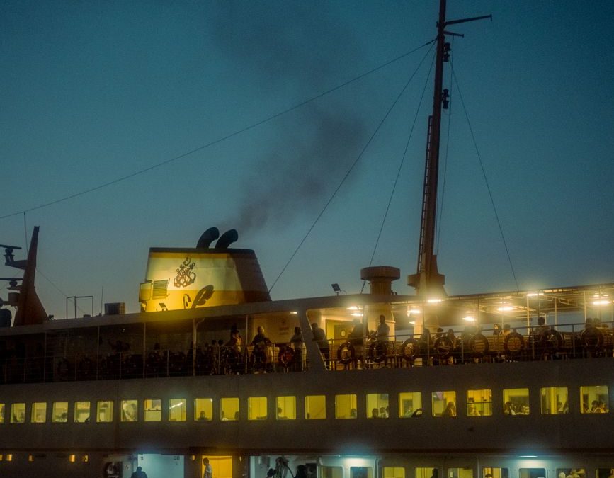 A ferry boat brightly lit, sailing at dusk with a reflection shimmering on the calm water.