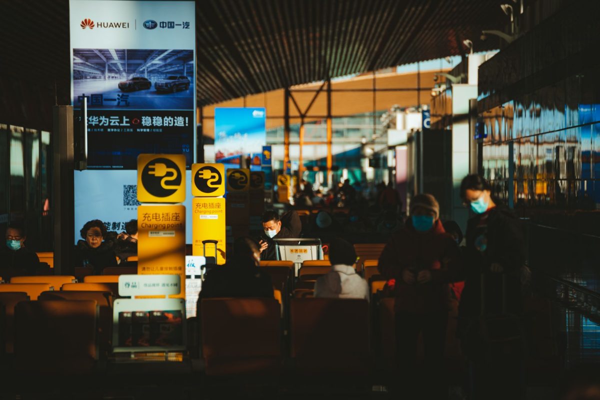 Crowded airport terminal scene with passengers wearing face masks and signage in multiple languages.
